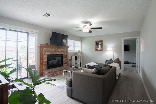 living area with ceiling fan, a textured ceiling, visible vents, a brick fireplace, and wood finished floors