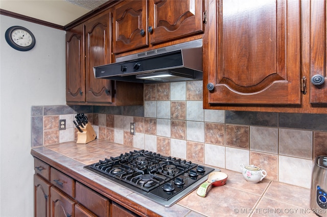 kitchen featuring crown molding, decorative backsplash, tile counters, under cabinet range hood, and black gas cooktop
