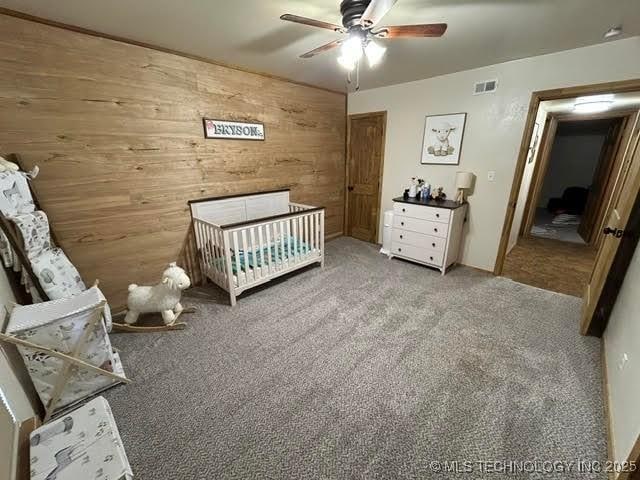 bedroom with wooden walls, ceiling fan, a crib, carpet floors, and visible vents