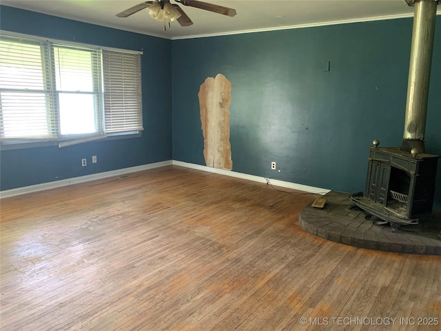 unfurnished living room with wood-type flooring, a wood stove, ornamental molding, baseboards, and a ceiling fan