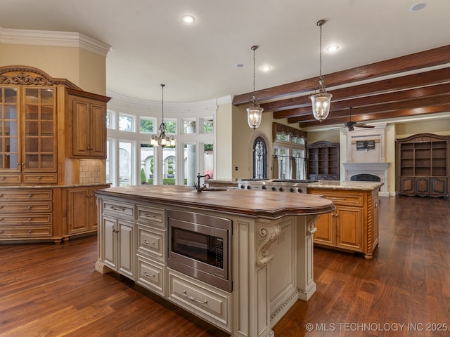 kitchen featuring a fireplace, wooden counters, an island with sink, ceiling fan with notable chandelier, and stainless steel microwave