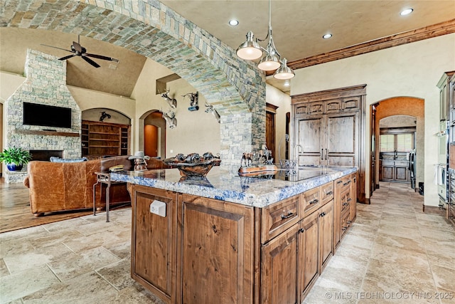 kitchen featuring stone tile floors, a center island with sink, arched walkways, light stone counters, and a ceiling fan