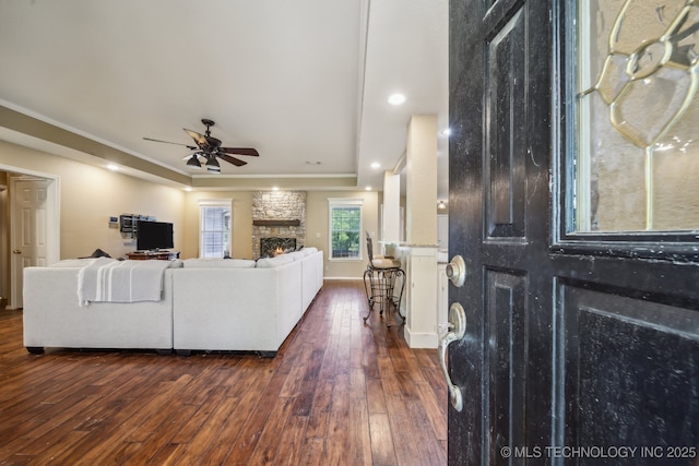 living room with crown molding, a ceiling fan, dark wood finished floors, and a stone fireplace