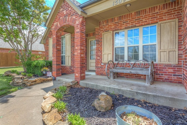 view of exterior entry with a porch, brick siding, and fence