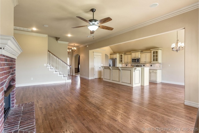 unfurnished living room with ceiling fan with notable chandelier, ornamental molding, a brick fireplace, arched walkways, and baseboards