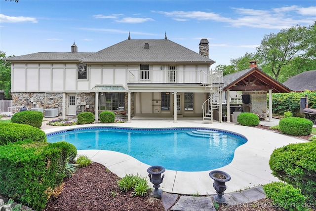 back of house featuring a balcony, a patio area, stone siding, a chimney, and stairs