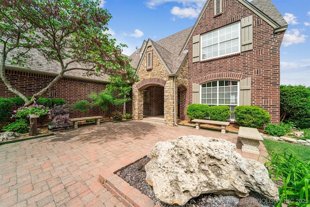 tudor-style house with brick siding, roof with shingles, and a patio
