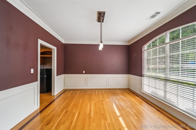 empty room with crown molding, light wood-style flooring, visible vents, and a wainscoted wall