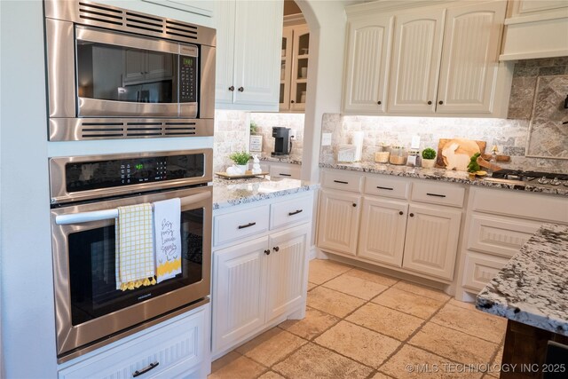 kitchen featuring stainless steel appliances, decorative backsplash, light stone countertops, arched walkways, and glass insert cabinets