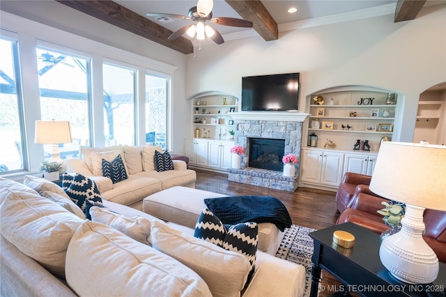living room featuring beamed ceiling, a ceiling fan, dark wood finished floors, a stone fireplace, and built in features