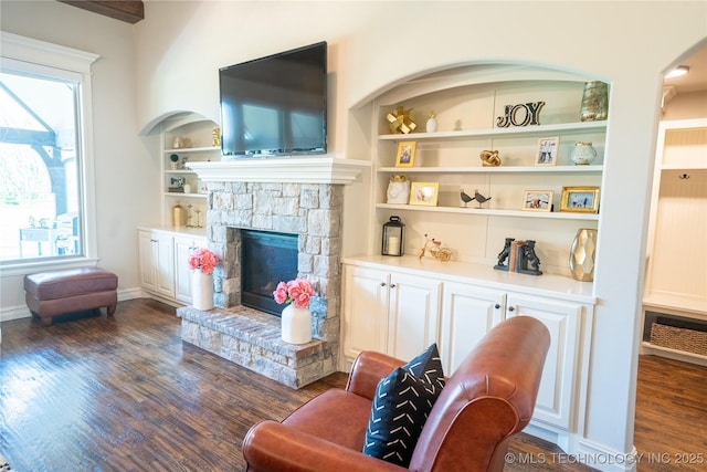 living room featuring dark wood-type flooring, a stone fireplace, built in shelves, and baseboards