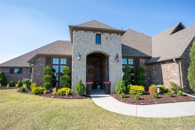 french provincial home featuring a shingled roof, a front lawn, brick siding, and stone siding