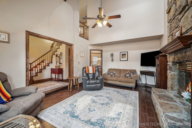 living area featuring wood finish floors, stairway, a ceiling fan, a towering ceiling, and a stone fireplace
