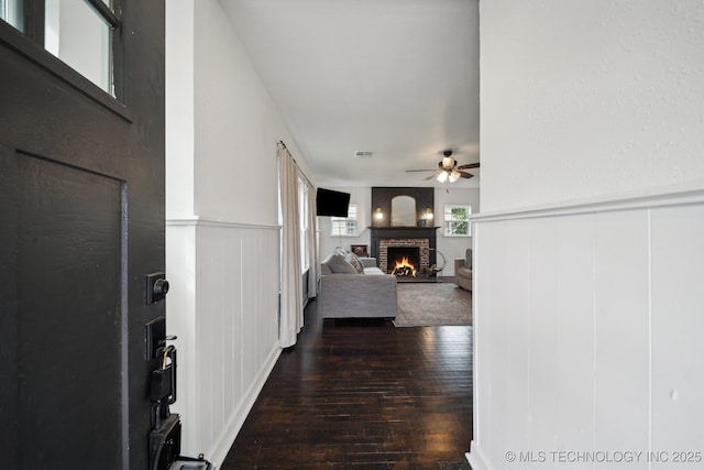 foyer featuring dark wood finished floors, ceiling fan, a brick fireplace, wainscoting, and a decorative wall