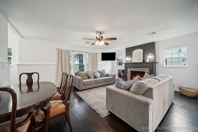 living area with dark wood-style flooring, plenty of natural light, a lit fireplace, ceiling fan, and a decorative wall