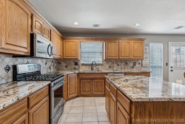 kitchen with appliances with stainless steel finishes, a sink, light stone counters, brown cabinets, and a textured ceiling