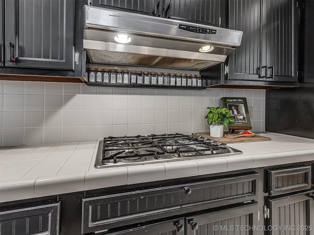 kitchen with tile counters, under cabinet range hood, oven, tasteful backsplash, and stainless steel gas stovetop