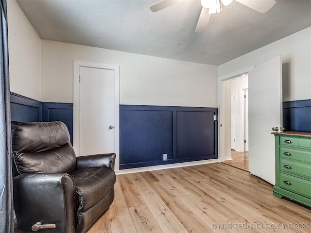 sitting room featuring a ceiling fan, a decorative wall, light wood-style floors, a wainscoted wall, and a textured ceiling