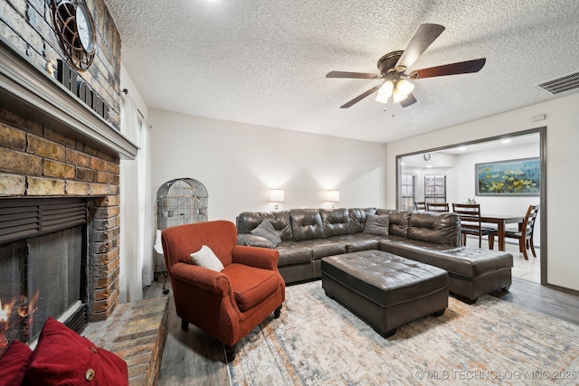 living area with wood finished floors, a ceiling fan, a textured ceiling, and a brick fireplace