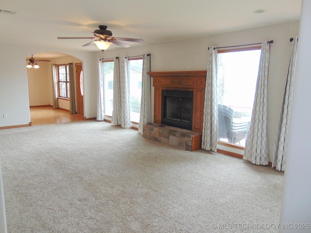 unfurnished living room featuring a ceiling fan, carpet floors, a fireplace with raised hearth, arched walkways, and baseboards