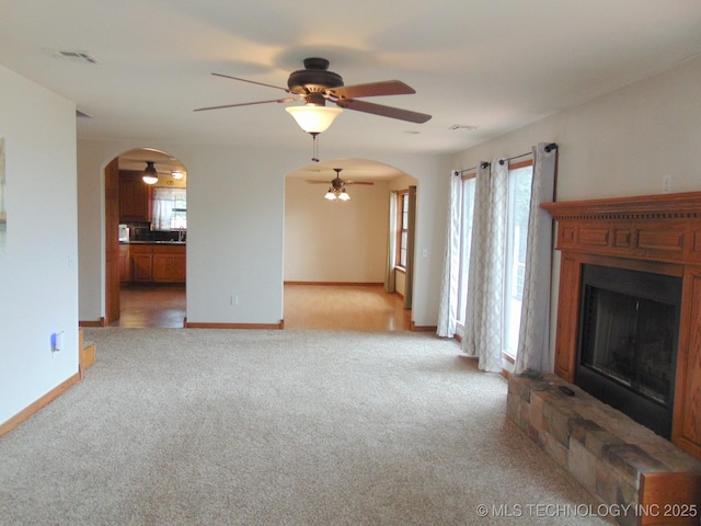 unfurnished living room featuring arched walkways, ceiling fan, a fireplace with raised hearth, carpet flooring, and baseboards