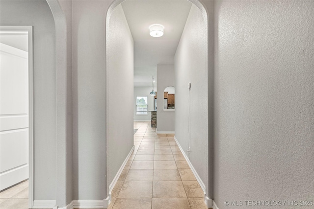 hall featuring light tile patterned flooring, arched walkways, a textured wall, and baseboards