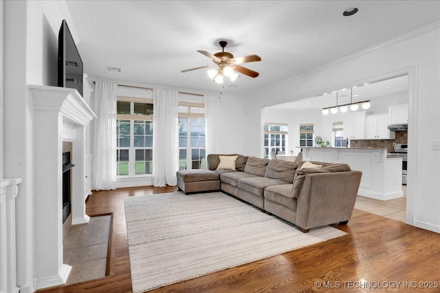 living area featuring a ceiling fan, a fireplace with flush hearth, ornamental molding, and light wood-style floors