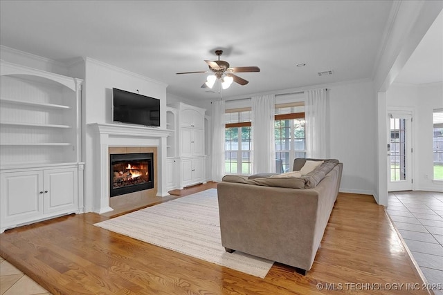 living area featuring a tiled fireplace, a ceiling fan, ornamental molding, built in features, and wood finished floors
