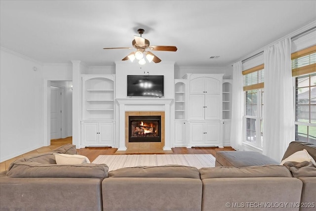 living area with a ceiling fan, a tile fireplace, built in shelves, light wood-type flooring, and ornamental molding