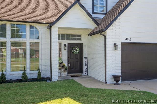 entrance to property featuring a shingled roof, brick siding, and a yard