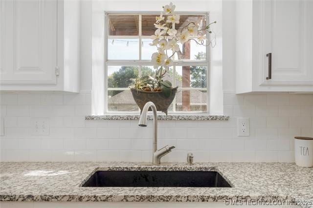 kitchen view of a sink, white cabinetry, light stone countertops, and backsplash