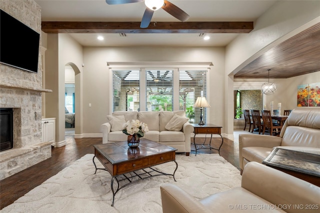 living area featuring arched walkways, wood finished floors, a ceiling fan, a stone fireplace, and beam ceiling