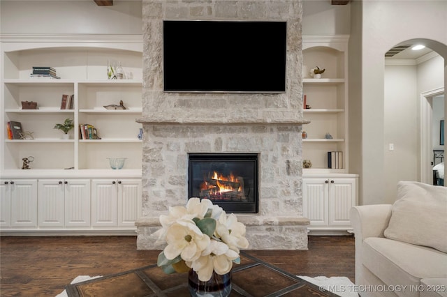 living room featuring arched walkways, dark wood-type flooring, a stone fireplace, built in features, and crown molding