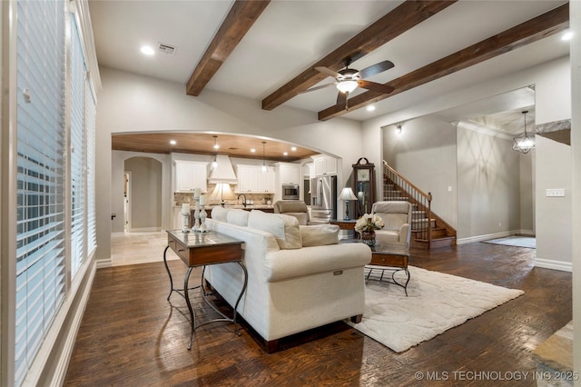 living area with arched walkways, ceiling fan, dark wood-type flooring, beam ceiling, and baseboards
