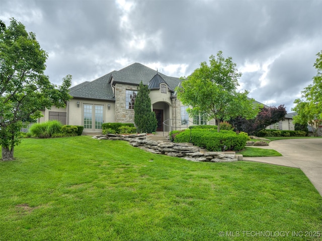 french country inspired facade featuring stone siding, french doors, a front yard, roof with shingles, and stucco siding
