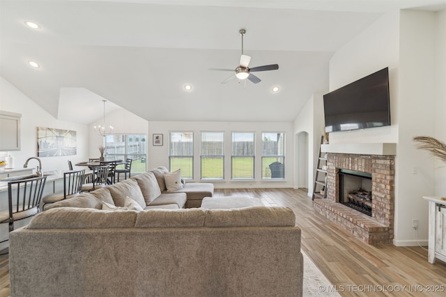 living area with vaulted ceiling, ceiling fan, light wood-type flooring, a brick fireplace, and a chandelier