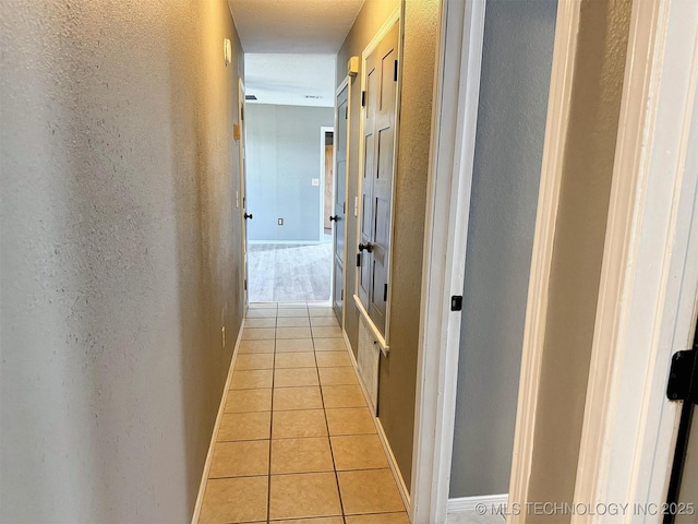corridor featuring light tile patterned floors, a textured wall, and baseboards