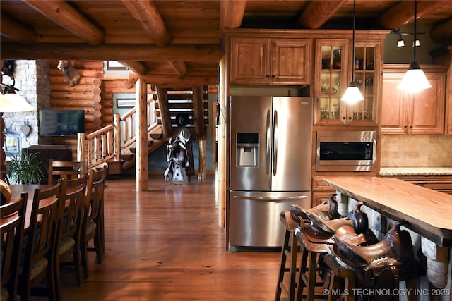 kitchen with stainless steel appliances, rustic walls, brown cabinetry, hardwood / wood-style flooring, and beamed ceiling