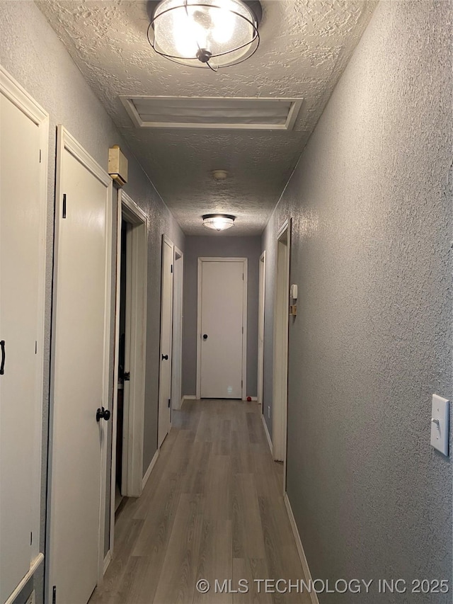 hall with attic access, wood finished floors, a textured wall, a textured ceiling, and baseboards