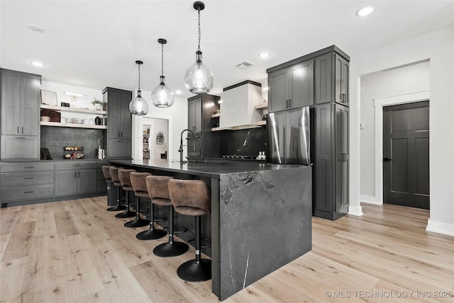 kitchen with open shelves, dark countertops, freestanding refrigerator, light wood-style floors, and a center island with sink