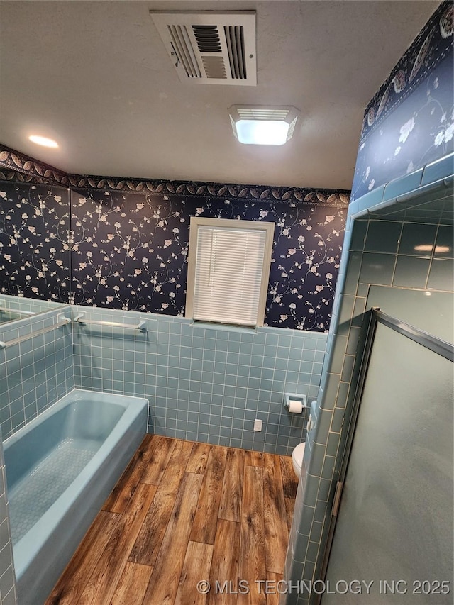 bathroom featuring wallpapered walls, wood finished floors, tile walls, a wainscoted wall, and a bath