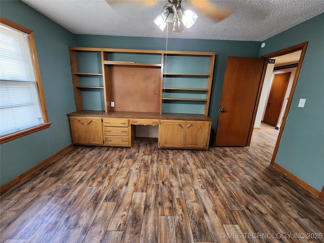 unfurnished office with built in study area, a textured ceiling, dark wood-style floors, ceiling fan, and baseboards