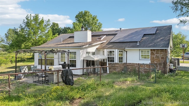 back of house featuring solar panels, a chimney, a patio area, fence, and a shingled roof