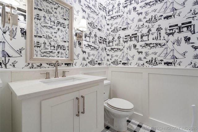 bathroom with wallpapered walls, a wainscoted wall, toilet, vanity, and a decorative wall