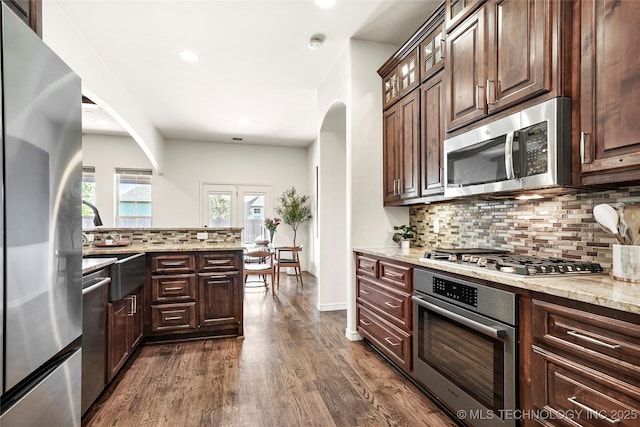 kitchen with appliances with stainless steel finishes, decorative backsplash, arched walkways, light stone counters, and dark wood-style floors