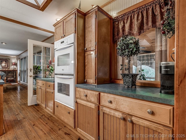 kitchen with white double oven, dark countertops, wood-type flooring, brown cabinetry, and ornamental molding
