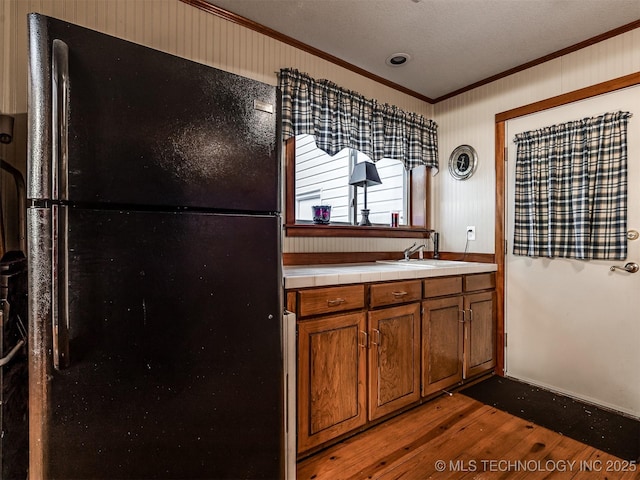 kitchen featuring freestanding refrigerator, crown molding, tile countertops, wood finished floors, and brown cabinets