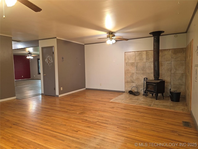 unfurnished living room featuring a ceiling fan, a wood stove, wood finished floors, crown molding, and baseboards