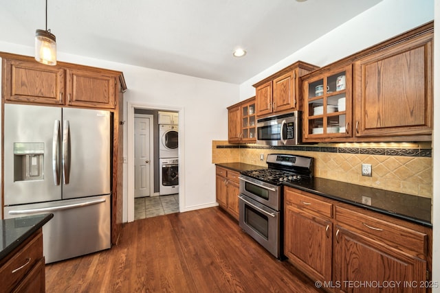 kitchen featuring stainless steel appliances, stacked washer and clothes dryer, brown cabinets, glass insert cabinets, and backsplash