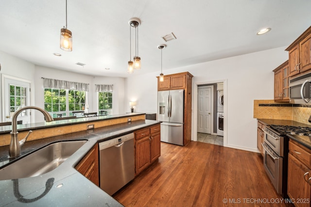 kitchen with stainless steel appliances, stacked washer / dryer, a sink, brown cabinets, and dark wood finished floors